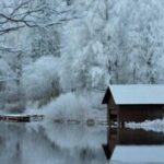 Eine kleine Holzhütte steht an einem ruhigen, spiegelnden See, umgeben von schneebedeckten Bäumen und Ästen in einer Winterlandschaft.