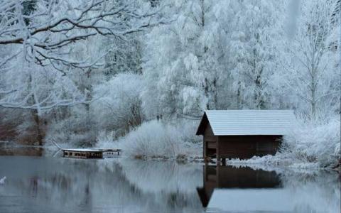 Eine kleine Holzhütte steht an einem ruhigen, spiegelnden See, umgeben von schneebedeckten Bäumen und Ästen in einer Winterlandschaft.
