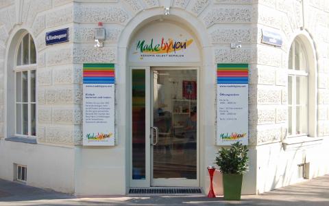 Schaufenster mit einer Glastür unter einem Bogenfenster, flankiert von bunten Schildern mit der Aufschrift "Made by You", an einem weißen Backsteingebäude an einer Straßenecke.