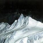 Ein schneebedeckter Berggipfel hebt sich scharf von einem dunklen Hintergrund ab, umgeben von zerklüftetem, eisigem Terrain.