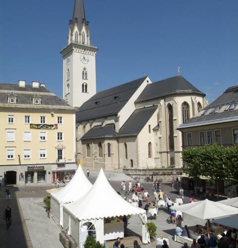 Ein Stadtplatz mit weißen Zelten, die für eine Veranstaltung im Freien aufgestellt sind, Menschen, die um Tische versammelt sind, und eine Kirche mit einem hohen Kirchturm im Hintergrund.