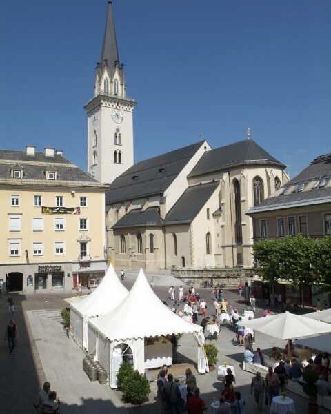 Ein Stadtplatz mit weißen Zelten, die für eine Veranstaltung im Freien aufgestellt sind, Menschen, die um Tische versammelt sind, und eine Kirche mit einem hohen Kirchturm im Hintergrund.