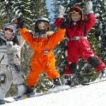 Drei Kinder in bunten Schneeanzügen stehen mit erhobenen Armen auf einem verschneiten Hang, umgeben von schneebedeckten Bäumen.