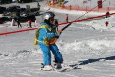 Ein Kind in blauer Skibekleidung und mit Helm benutzt auf einer verschneiten Piste einen Schlepplift und hält sich am Seil fest, während es auf Skiern steht.