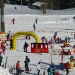 Kinder und Erwachsene fahren Ski und spielen in einem verschneiten Gebiet mit farbenfrohen Dekorationen, einer roten Zugstruktur und verschiedenen Schildern. Im Hintergrund sind schneebedeckte Bäume zu sehen.