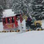 Ein roter Passagierschlitten wird von einem gelben Schneemobil durch einen verschneiten Wald mit schneebedeckten Bäumen im Hintergrund gezogen.
