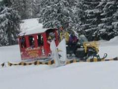 Ein roter Passagierschlitten wird von einem gelben Schneemobil durch einen verschneiten Wald mit schneebedeckten Bäumen im Hintergrund gezogen.