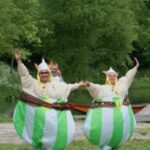 Zwei Personen in grünen und weißen aufblasbaren Kostümen halten sich an den Händen und posieren mit erhobenen Armen im Freien in der Nähe eines Sees, wobei eine dritte Person hinter ihnen steht. Im Hintergrund sind Bäume und ein Auto zu sehen.