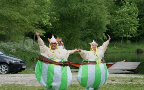 Zwei Personen in grünen und weißen aufblasbaren Kostümen halten sich an den Händen und posieren mit erhobenen Armen im Freien in der Nähe eines Sees, wobei eine dritte Person hinter ihnen steht. Im Hintergrund sind Bäume und ein Auto zu sehen.