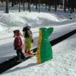 Drei Kinder in Winterkleidung, eines davon in einem grünen Dinosaurierkostüm, stehen auf einem Förderbandlift in einem verschneiten Waldgebiet.