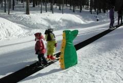Drei Kinder in Winterkleidung, eines davon in einem grünen Dinosaurierkostüm, stehen auf einem Förderbandlift in einem verschneiten Waldgebiet.