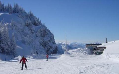 Zwei Personen fahren auf einer verschneiten Bergpiste unter strahlend blauem Himmel Ski, im Hintergrund sind schneebedeckte Bäume und Gebäude zu sehen.
