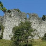 Eine teilweise zerstörte steinerne Burgmauer steht auf einem grasbewachsenen Hügel, mit Bäumen und Büschen im Vordergrund unter einem klaren blauen Himmel.
