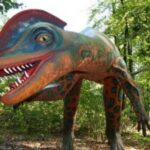 Eine farbenfrohe, realistische Dinosaurier-Statue mit scharfen Zähnen steht in einem Waldgebiet, umgeben von grünen Bäumen und Laub.