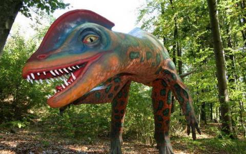 Eine farbenfrohe, realistische Dinosaurier-Statue mit scharfen Zähnen steht in einem Waldgebiet, umgeben von grünen Bäumen und Laub.