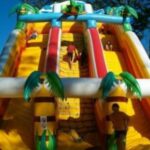 Kinder klettern und spielen auf einer großen, aufblasbaren Dschungelrutsche mit Palmendekoration.