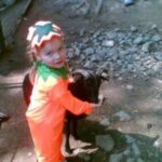 Ein kleines Kind in einem orangefarbenen Kürbiskostüm streichelt eine schwarze Ziege im Freien auf einem schmutzigen und felsigen Boden in der Nähe eines eingezäunten Geländes.