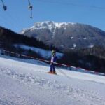 Eine Person in Skiausrüstung steht auf einer verschneiten Piste mit Bergen und Bäumen im Hintergrund, getrennt durch ein rot-weißes Absperrband.