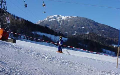 Eine Person in Skiausrüstung steht auf einer verschneiten Piste mit Bergen und Bäumen im Hintergrund, getrennt durch ein rot-weißes Absperrband.