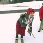 Eine Person in traditioneller Kleidung mit Kopftuch und Holzschuhen steht mit einem Stock in der Hand im Schnee, eine weitere Person ist teilweise im Hintergrund zu sehen.