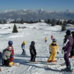 Eine Gruppe von Kindern in Winterkleidung steht auf einer verschneiten Skipiste mit Bergen und Bäumen im Hintergrund.