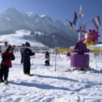 Menschen stehen in einer verschneiten Gegend in der Nähe eines bunten Karussells, mit Bergen und klarem Himmel im Hintergrund.