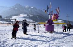 Menschen stehen in einer verschneiten Gegend in der Nähe eines bunten Karussells, mit Bergen und klarem Himmel im Hintergrund.