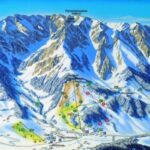 Illustrierte Skigebietskarte mit schneebedeckten Berggipfeln, mit farbigen Linien markierten Skipisten und Liftrouten im Vordergrund.