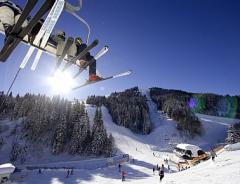 An einem sonnigen Tag fahren Skifahrer mit einem Sessellift über einen verschneiten Berghang, unter dem Bäume, Skifahrer und eine Skihütte zu sehen sind.