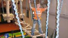 Ein Kind, das einen Sicherheitsgurt trägt, läuft auf einer Hängeseilbrücke in einem Indoor-Abenteuerspielplatz.