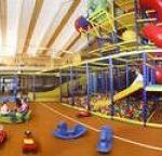 Indoor-Spielplatz mit einem großen Klettergerüst, Rutschen, einer Ballgrube und verschiedenen Plastikspielzeugen auf einem braunen Boden. Mehrere Kinder spielen in dem Bereich.