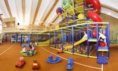 Indoor-Spielplatz mit einem großen Klettergerüst, Rutschen, einer Ballgrube und verschiedenen Plastikspielzeugen auf einem braunen Boden. Mehrere Kinder spielen in dem Bereich.