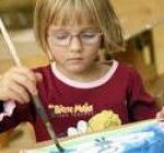 Ein kleines Kind mit Brille malt an einem Tisch mit einem Pinsel auf Papier und trägt ein rotes Hemd mit Zeichentrickfiguren.