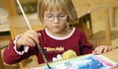 Ein kleines Kind mit Brille malt an einem Tisch mit einem Pinsel auf Papier und trägt ein rotes Hemd mit Zeichentrickfiguren.