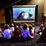 Eine Gruppe von Kindern sitzt in einem schwach beleuchteten Raum auf dem Boden und sieht sich einen auf eine große Leinwand projizierten Film an.