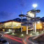 Ein modernes Gebäude mit großen Glasfenstern und einem hohen Wasserrutschenkomplex im Freien in der Abenddämmerung, mit einigen Bergen im Hintergrund und Auto-Leuchtpfaden davor.