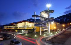 Ein modernes Gebäude mit großen Glasfenstern und einem hohen Wasserrutschenkomplex im Freien in der Abenddämmerung, mit einigen Bergen im Hintergrund und Auto-Leuchtpfaden davor.