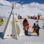 Kinder und ein Erwachsener stehen neben einem weißen Tipi auf einer verschneiten Skipiste, mit bunten Dekorationen und verschneiten Bergen im Hintergrund.