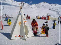 Kinder und ein Erwachsener stehen neben einem weißen Tipi auf einer verschneiten Skipiste, mit bunten Dekorationen und verschneiten Bergen im Hintergrund.