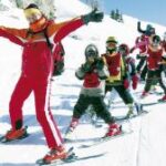 Ein Skilehrer in roter Kleidung führt eine Gruppe von Kindern in farbenfrohen Outfits auf einer verschneiten Piste an, die alle Skier und Winterkleidung tragen.