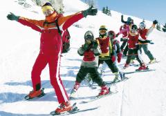 Ein Skilehrer in roter Kleidung führt eine Gruppe von Kindern in farbenfrohen Outfits auf einer verschneiten Piste an, die alle Skier und Winterkleidung tragen.