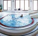 Indoor-Spa-Bereich mit Jacuzzi-Pool, großen Fenstern, weißen Wänden, einer Pflanze in der Ecke und einer Person im Wasser.