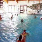 Menschen schwimmen und spielen in einem Hallenbad mit natürlichem Licht, das durch große Fenster einfällt; an der Wand im Hintergrund ist ein Wandgemälde zu sehen.