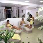 Drei Personen in Handtüchern entspannen sich auf Liegestühlen in einem Spa-ähnlichen Raum mit Pflanzen und gefliesten Böden.