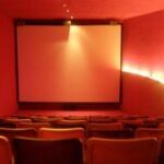 Ein kleiner, leerer Kinosaal mit roten Wänden, gelben Sitzreihen und einer leeren Leinwand im vorderen Bereich.