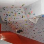 Eine Person klettert an einer Boulderwand in der Halle mit bunten Griffen und einer schrägen Decke; darunter ein rot gepolsterter Boden.
