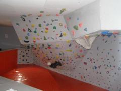Eine Person klettert an einer Boulderwand in der Halle mit bunten Griffen und einer schrägen Decke; darunter ein rot gepolsterter Boden.