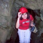 Eine Person, die einen roten Schutzhelm und ein rotes Hemd trägt, geht mit einer Taschenlampe in der Hand durch einen engen, felsigen Tunnel.