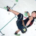 Eine Person in Kletterausrüstung erklimmt eine Indoor-Kletterwand, hält sich an grünen Griffen fest und lächelt in die Kamera.