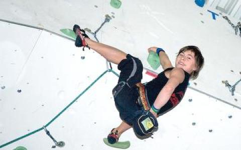 Eine Person in Kletterausrüstung erklimmt eine Indoor-Kletterwand, hält sich an grünen Griffen fest und lächelt in die Kamera.